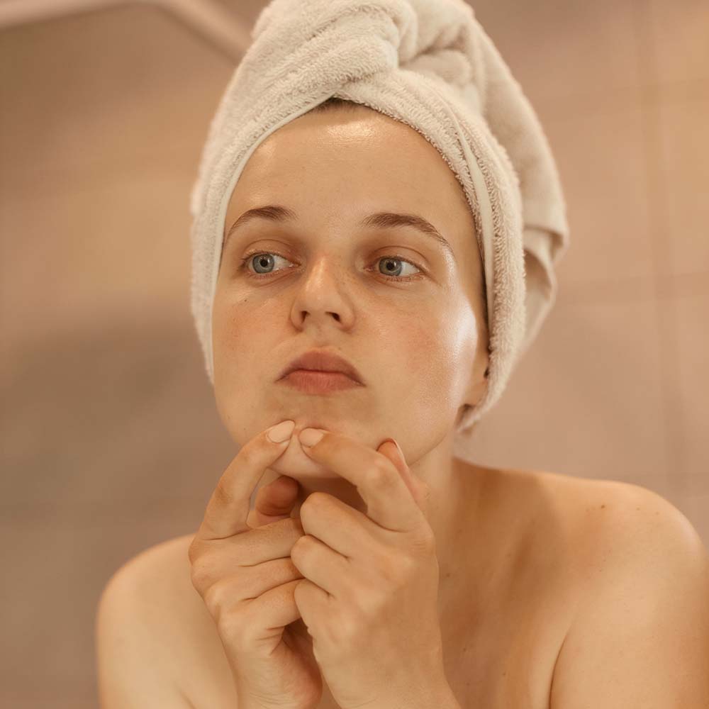  A woman with a white towel wrapped around her hair is squeezing a pimple on her nose with a displeased expression, exemplifying a common mistake to avoid in acne skincare routines for clearer skin.